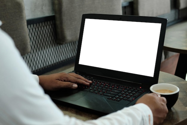 Mockup image of a man using and working on laptop computer with a blank white screen while drinking coffee on wooden table in cafe