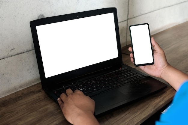 Макет изображения человека, использующего и работающего на ноутбуке и мобильном телефоне с пустым белым экраном на деревянном столе