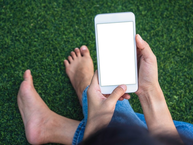 Mockup image of the phone. White blank screen on mobile phone in hand on green grass background. Women wearing white sleeveless shirt and jeans pant using smartphone with empty screens, top view.