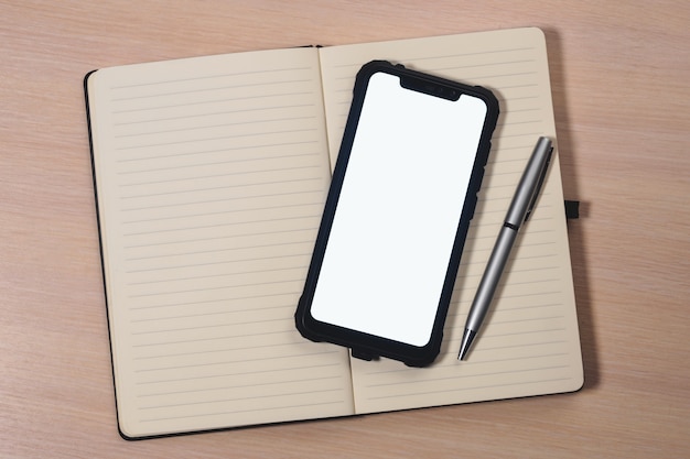 Mockup image of a smartphone with the blank white screen, pen and a notebook on a wooden table.