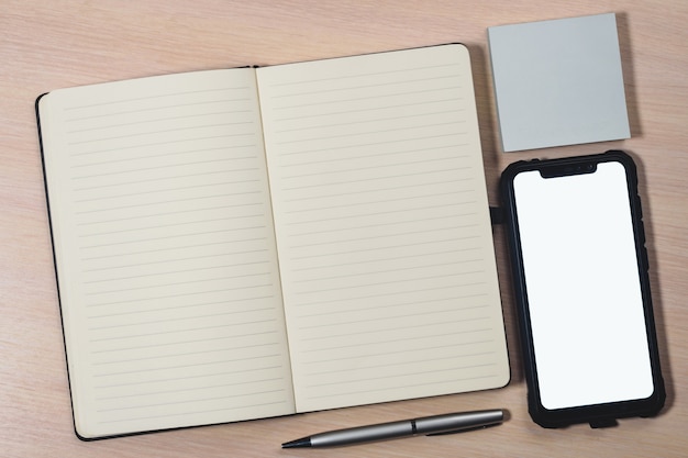 Mockup image of a smartphone with the blank white screen, pen, sticky notes and a notebook on a wooden table.