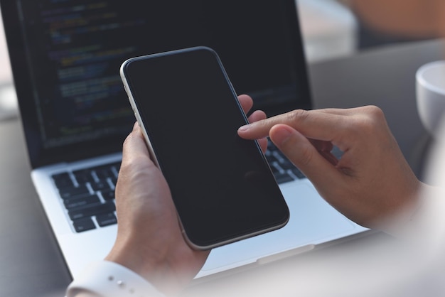 Mockup image of woman hand using blank screen mobile phone during working on laptop computer on office table