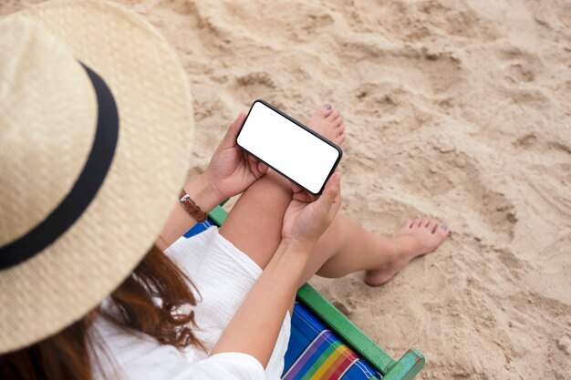 ビーチチェアに座っている間空白のデスクトップ画面で黒い携帯電話を保持している女性のモックアップ画像