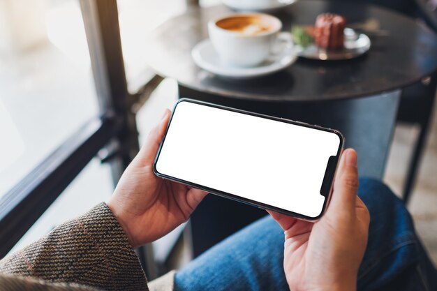 Mockup image of a woman holding black mobile phone with blank desktop screen while sitting in cafe