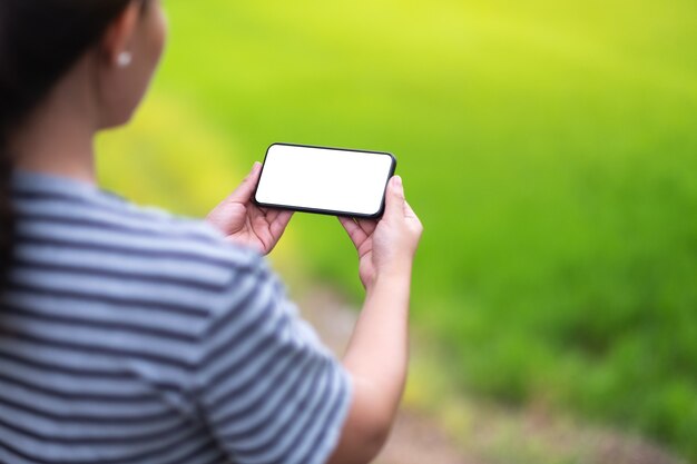 ぼやけた緑の自然の背景と空白のデスクトップ画面で黒の携帯電話を保持している女性のモックアップ画像