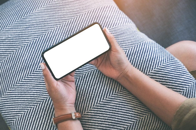 Mockup image of a woman holding black mobile phone with blank white desktop screen horizontally while sitting in living room with feeling relaxed