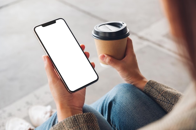 Mockup image of a woman holding black mobile phone with blank white screen while drinking coffee and sitting on the floor