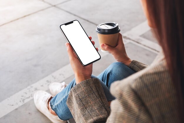 コーヒーを飲みながら床に座っている間、空白の白い画面で黒い携帯電話を保持している女性のモックアップ画像