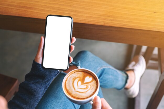 カフェに座っている間空白の白い画面で黒い携帯電話を保持している女性のモックアップ画像