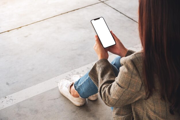 바닥에 앉아 있는 동안 빈 흰색 화면이 있는 검은색 휴대폰을 들고 있는 여성의 모형 이미지