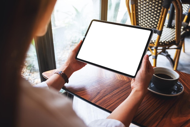 テーブルの上にコーヒーカップと空白の白い画面で黒いタブレットPCを保持している女性のモックアップ画像