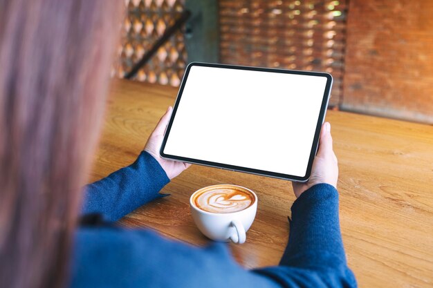 Mockup image of a woman holding black tablet pc with blank white screen with coffee cup on wooden table