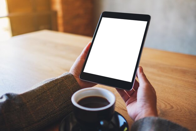 Mockup image of woman holding black tablet pc with blank white screen with coffee cup on wooden table