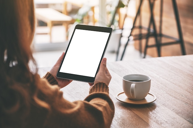 Mockup image of a woman holding black tablet pc with blank white screen with coffee cup on wooden table