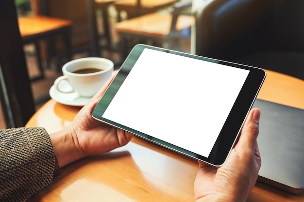 Mockup image of a woman holding black tablet pc with blank white screen with laptop and coffee cup on wooden table