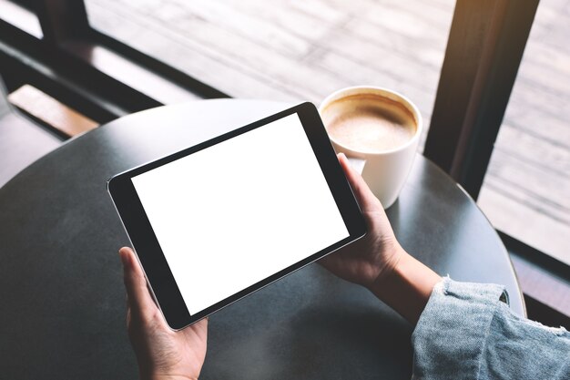 Mockup image of a woman holding black tablet with white blank screen and coffee cup on the table