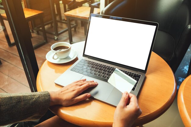 나무 테이블에 빈 흰색 화면과 커피 컵이 있는 노트북을 사용하는 동안 신용 카드를 들고 있는 여성의 흉내낸 이미지