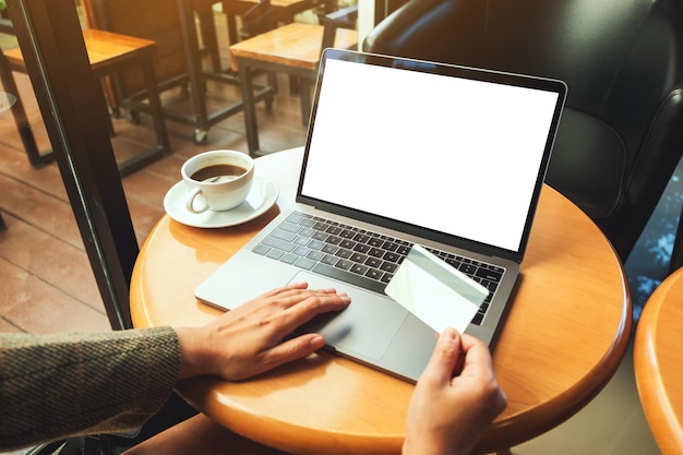 Immagine mockup di una donna in possesso di carta di credito mentre utilizza un laptop con schermo bianco vuoto e tazza di caffè su un tavolo di legno Foto Premium
