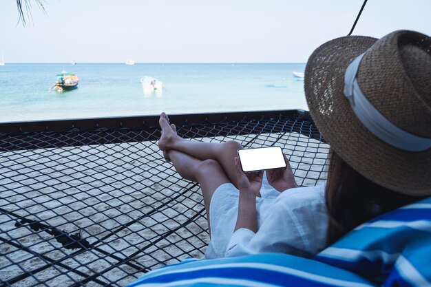 Макет изображения женщины, держащей мобильный телефон с пустым экраном рабочего стола, лежащей в гамаке на пляже