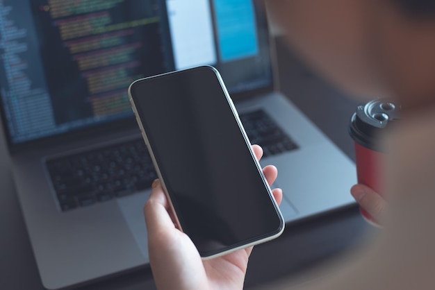 Mockup image woman holding mobile phone with blank screen template during working on laptop