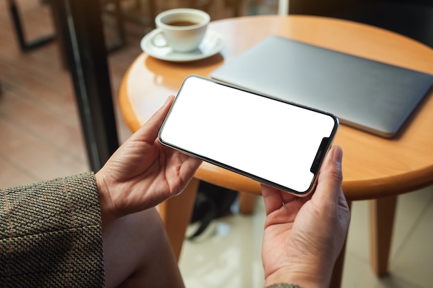 Mockup image of a woman holding mobile phone with blank white desktop screen with laptop and coffee cup on table