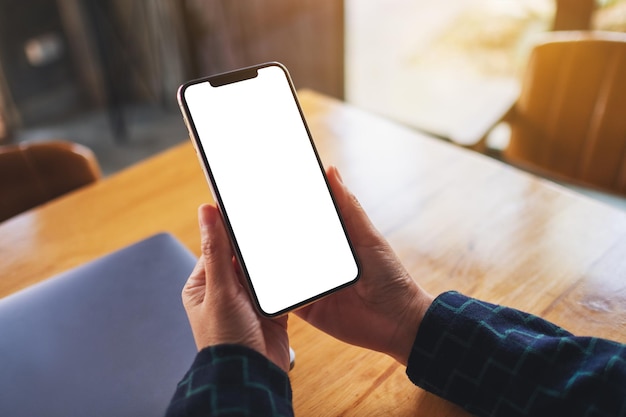 Mockup image of a woman holding mobile phone with blank white desktop screen with laptop on table