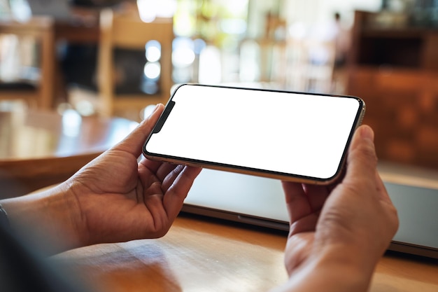 Mockup image of a woman holding mobile phone with blank white desktop screen with laptop on the table