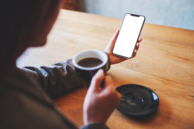 木製のテーブルでコーヒーを飲みながら空白の白い画面で携帯電話を保持している女性のモックアップ画像