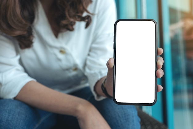 カフェで空白の白い画面で黒い携帯電話を持って見せている女性のモックアップ画像
