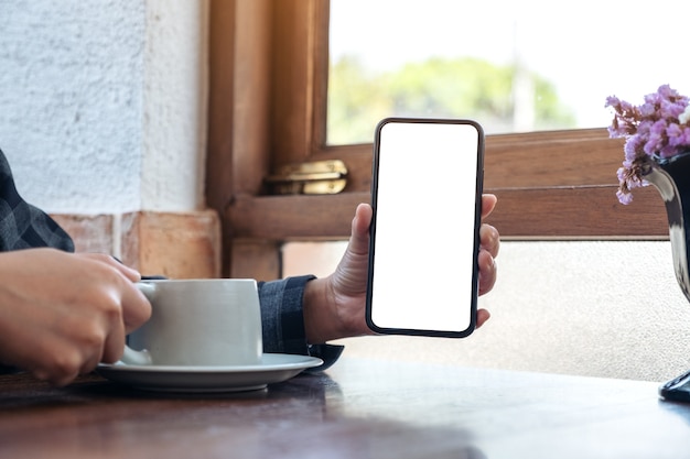 カフェでコーヒーを飲みながら、空白の白い画面で黒い携帯電話を持って見せている女性のモックアップ画像