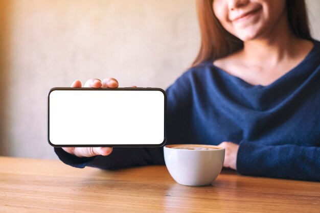 Mockup image of a woman holding and showing black mobile phone with blank white screen while drinking coffee