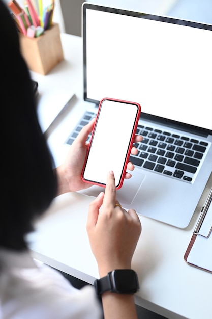 Mockup image of a woman holding smart phone with blank desktop screen while sitting in front of her laptop