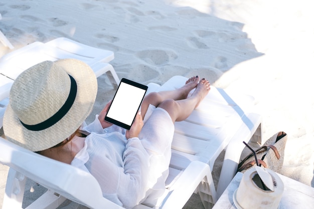 Mockup image of a woman holding and using a black tablet pc with blank desktop screen while laying down on beach chair on the beach