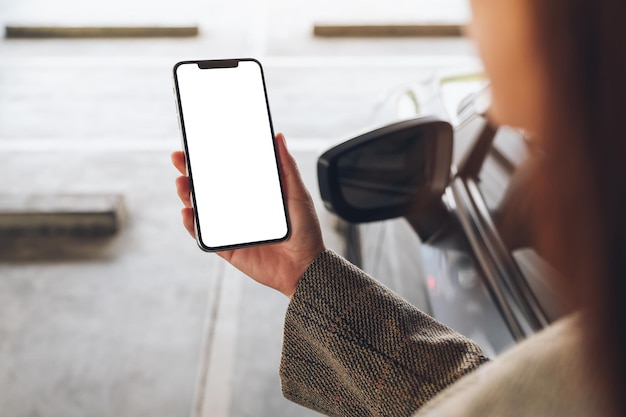 Photo mockup image of a woman holding and using mobile phone with blank white screen next to the car in parking lot