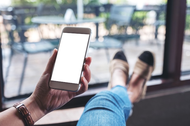 Photo mockup image of a woman holding white mobile phone with blank desktop screen while sitting in cafe