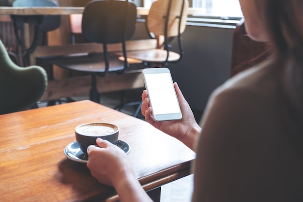 モダンなカフェでコーヒーを飲みながら空白の画面で白い携帯電話を保持している女性のモックアップ画像