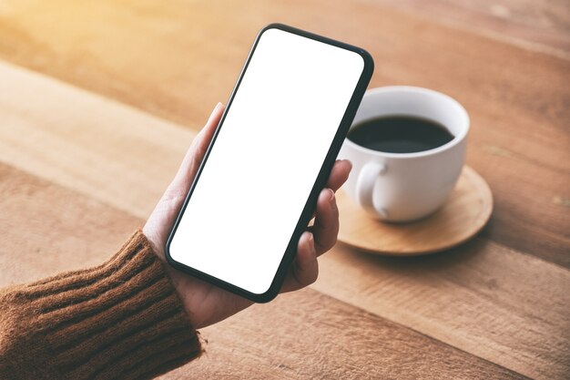 Immagine del modello della mano della donna che tiene il telefono cellulare nero con lo schermo in bianco con la tazza di caffè sulla tavola di legno