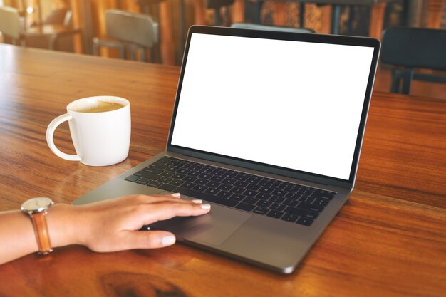 木製のテーブルにコーヒーカップと空白の白いデスクトップ画面でノートパソコンのタッチパッドを使用して触れている女性の手のモックアップ画像