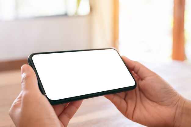 Mockup image of woman's hands holding black mobile phone with blank white screen horizontally on wooden table