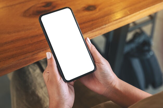 Mockup image of woman's hands holding black mobile phone with blank white screen on wooden table