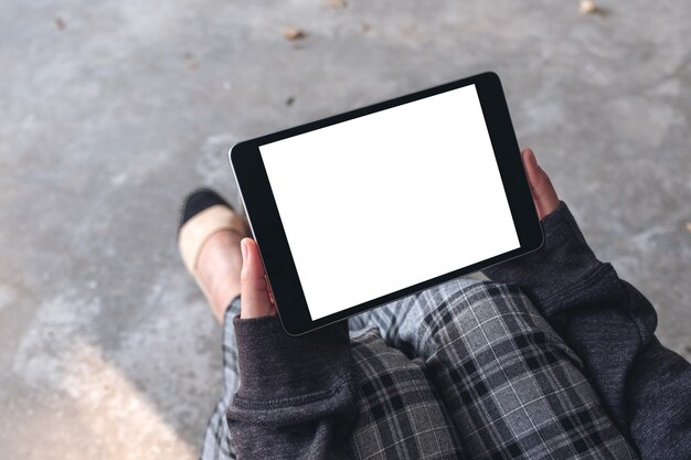 Mockup image of woman's hands holding and using black tablet pc with blank white desktop screen horizontally while sitting on the floor