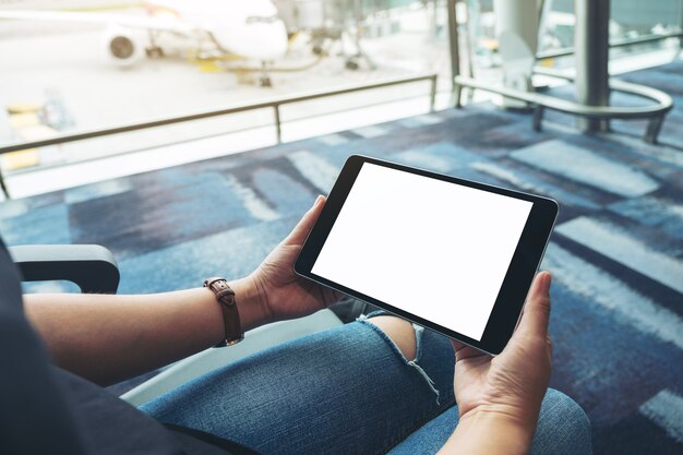 Immagine mockup delle mani di una donna che tiene e usa un tablet pc nero con schermo bianco vuoto mentre è seduta in aeroporto