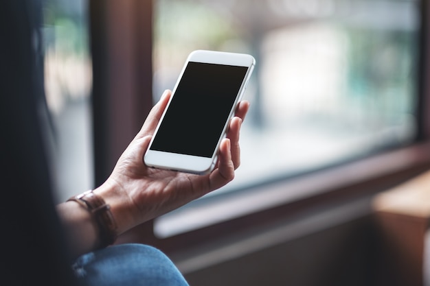 Mockup image of woman's hands holding white mobile phone with blank black desktop screen in cafe