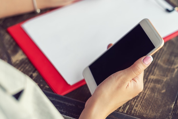 Mockup image of a woman's hands holding white mobile phone with blank black screen on background of white sheet of paper at a cafe.