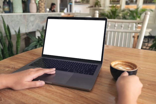 Immagine del modello delle mani della donna che digita e che tocca sul touchpad del computer portatile con lo schermo del desktop bianco vuoto mentre beve il caffè sulla tavola di legno nella caffetteria