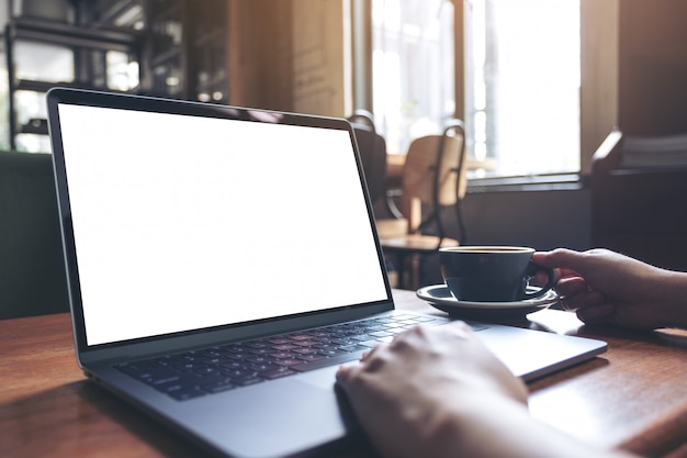 Mockup image of a woman using laptop with blank white desktop screen while drinking hot coffee on wooden table in cafe