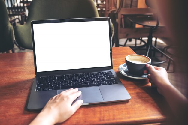 Mockup image of a woman using laptop with blank white desktop screen while drinking hot coffee on wooden table in cafe