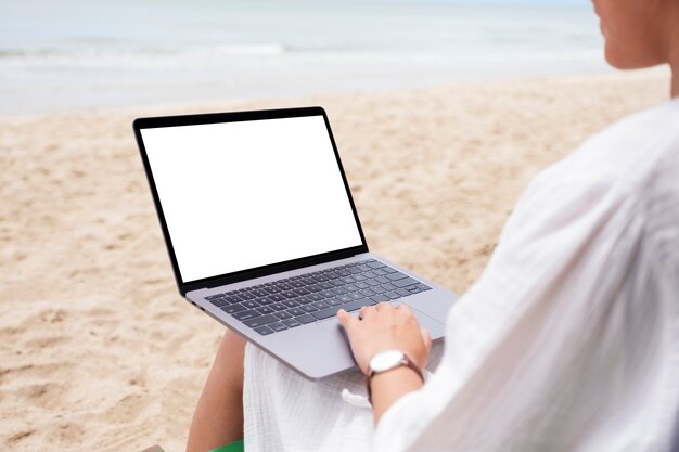 해변 의자에 앉아 빈 데스크탑 화면이 있는 랩톱 컴퓨터를 사용하고 타이핑하는 여성의 모형 이미지