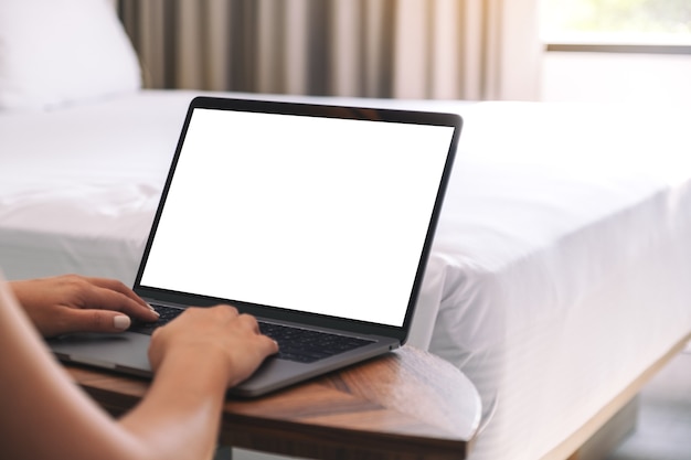 Mockup image of a woman using and typing on laptop with blank white desktop screen keyboard on wooden table next to the bed