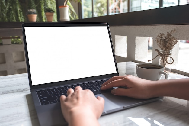 Mockup image of a woman using and typing on laptop with blank white screen and coffee cup on table in modern cafe